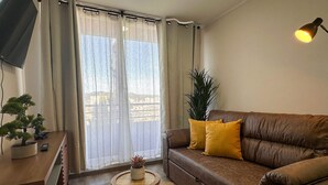 A living room with a brown leather sofa, a small wooden side table, a potted plant, and a large window with curtains. - Vista Laguna Oriente By Andes+ (Concepción)
