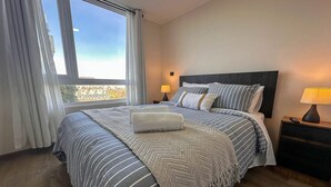 A hotel room with a large bed, a nightstand with a lamp, and a view of the city through the window. - Vista Laguna Oriente By Andes+ (Concepción)