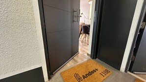 A hallway with a dark door, a doormat with a name and number, and a glimpse of a dining area with chairs and a table. - Vista Laguna Oriente By Andes+ (Concepción)