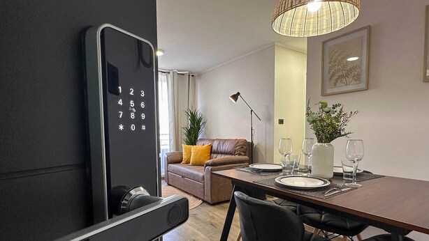 A modern living room with a dining area, featuring a large black smartphone on a stand, a brown leather sofa, a dining table set with plates, glasses, and a vase of flowers. - Vista Laguna Oriente By Andes+ (Concepción)
