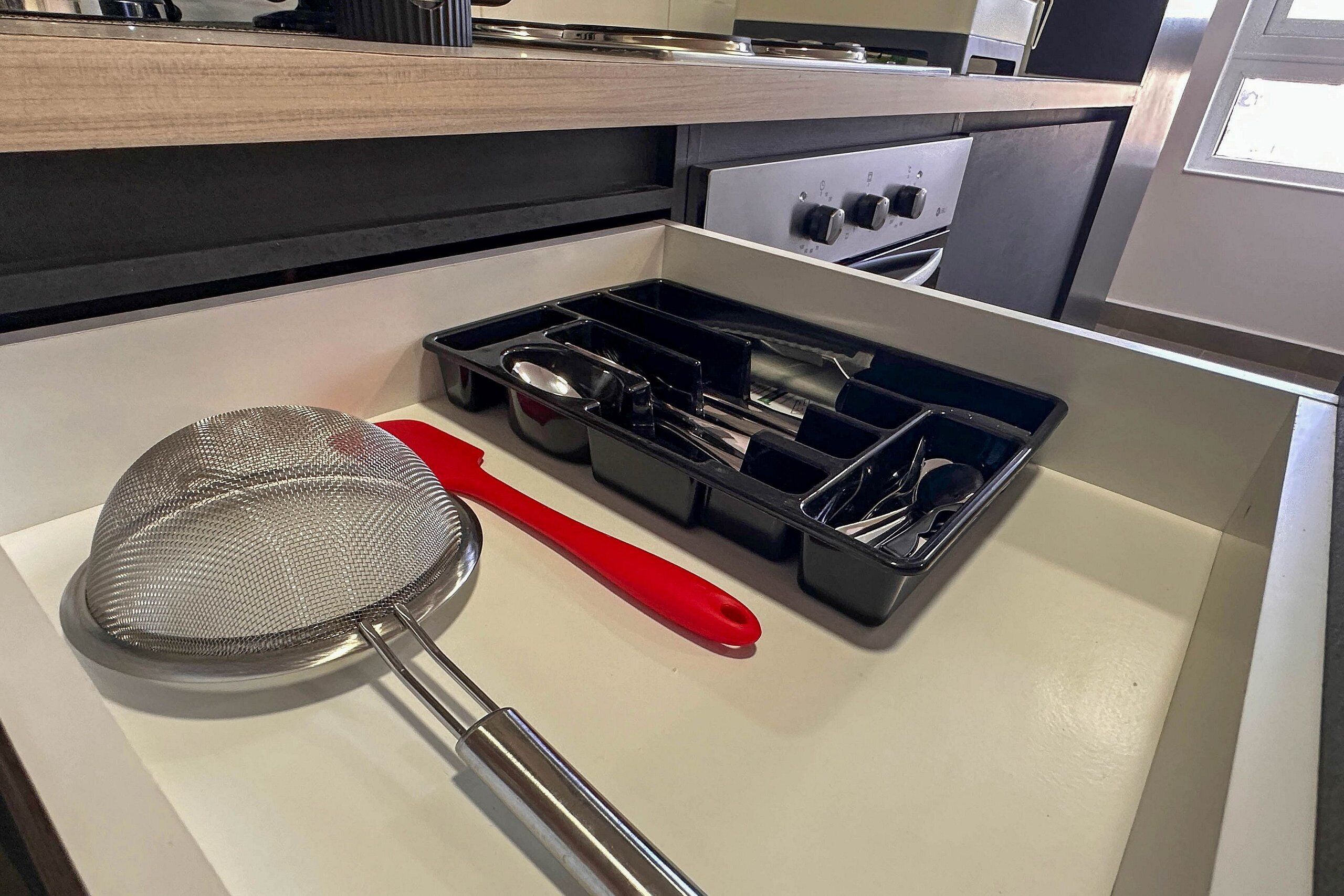 Un cajón de cocina con un colador, una espátula roja y un juego de utensilios negros.