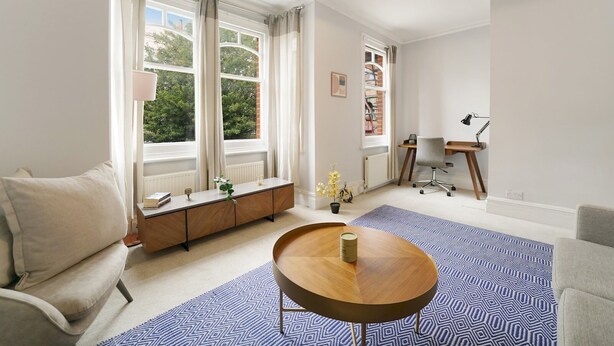 A modern living room with a sofa, a round coffee table, and a desk with a chair. There are large windows with curtains, a radiator, and a bookshelf. - Pass the Keys | Cosy apartment in Kilburn (London)