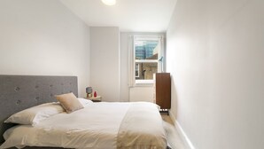 A bedroom with a grey upholstered headboard, a bed with white linens, a small bedside table with a lamp, and a view of the outside through a window. - Pass the Keys | Cosy apartment in Kilburn (London)