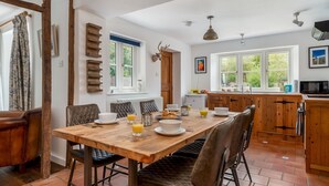 A dining area with a wooden table set for breakfast, surrounded by leather chairs. A kitchen with wooden cabinets and a window is visible in the background. - Pass the Keys | Cotswold Country Style Living | Chipping Norton (Chipping Norton)