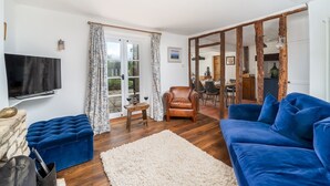 A living room with a blue sofa, a wooden coffee table, a TV mounted on the wall, and a large window with curtains. - Pass the Keys | Cotswold Country Style Living | Chipping Norton (Chipping Norton)