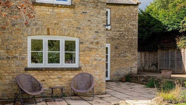 A stone building with two windows and a door, a small outdoor seating area with two chairs and a table, and a garden with a wooden fence and plants. - Pass the Keys | Cotswold Country Style Living | Chipping Norton (Chipping Norton)