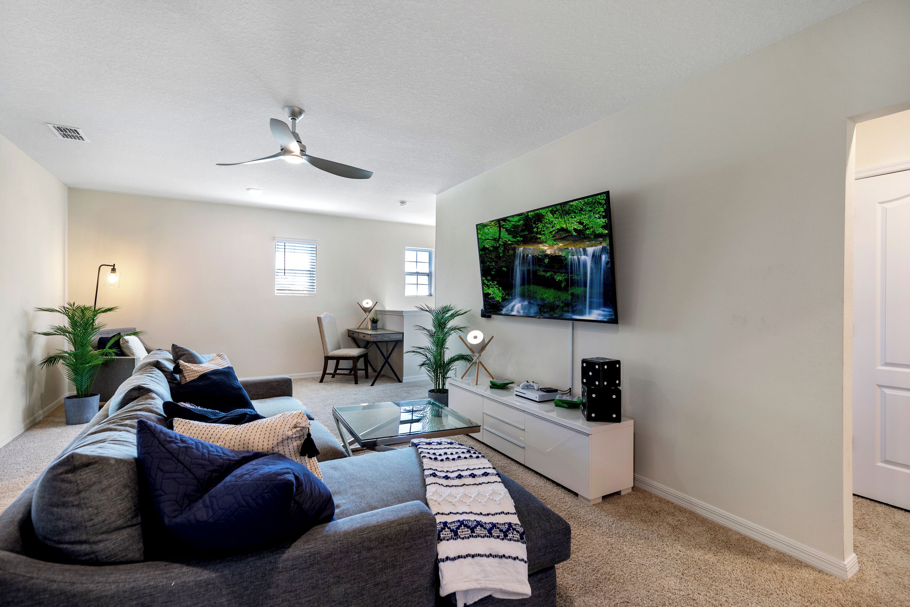 A modern living room with a gray sofa, a glass coffee table, a flat-screen TV mounted on the wall, and a ceiling fan.