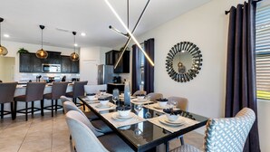 A modern dining area with a long table set for six, surrounded by grey upholstered chairs. A kitchen with dark wooden cabinets and a central island is visible in the background. - Solterra- LakeView - Private Pool & Spa  (Davenport)