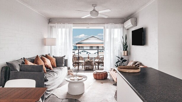 A modern living room with a sofa, coffee table, and dining table. There is a ceiling fan, a TV mounted on the wall, and a view of the outside balcony. - Pacific Sun CBD Apartment (Yamba)