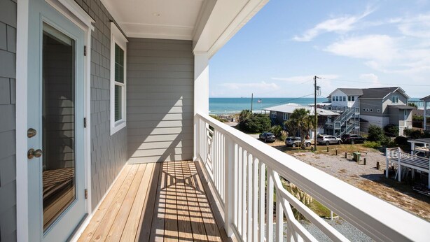 Un balcon doté d’une terrasse en bois, de balustrades blanches et offrant une vue sur l’océan et les maisons voisines.