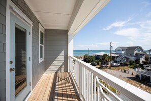 A balcony with a wooden deck, white railings, and a view of the ocean and neighboring houses.