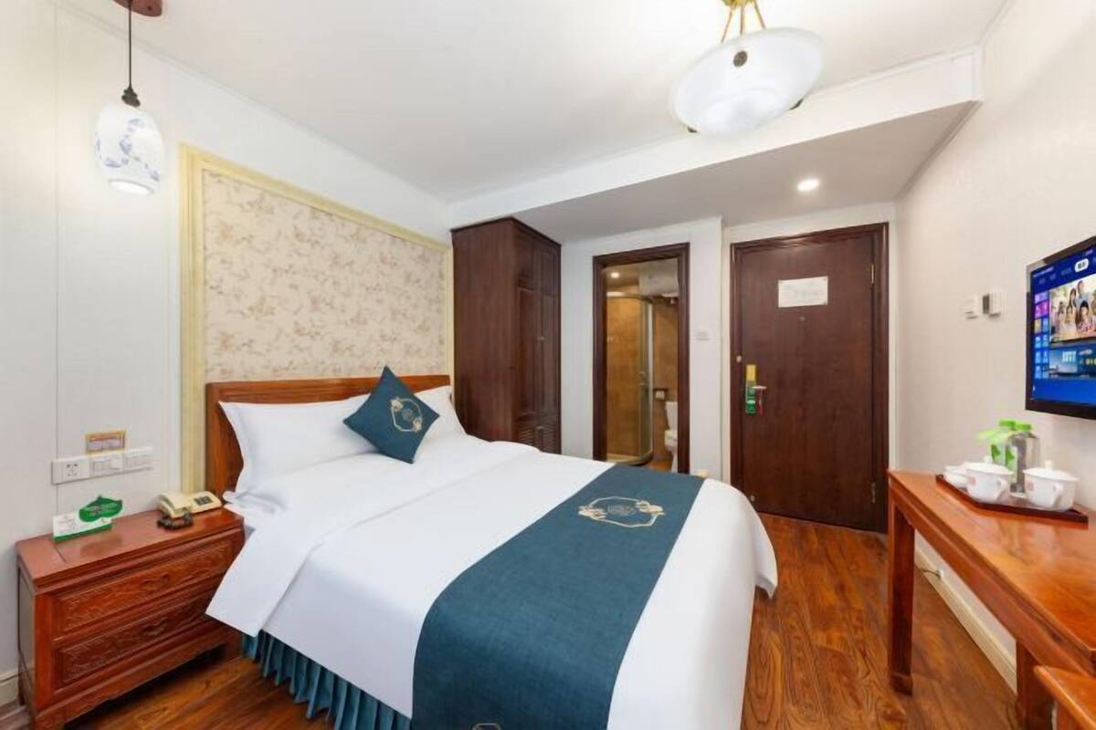 Una habitación de hotel con cama, mesita de noche, lámpara y televisión en la pared.