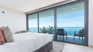 A bedroom with a large bed, a view of the ocean through floor-to-ceiling glass doors, and a seating area with chairs.