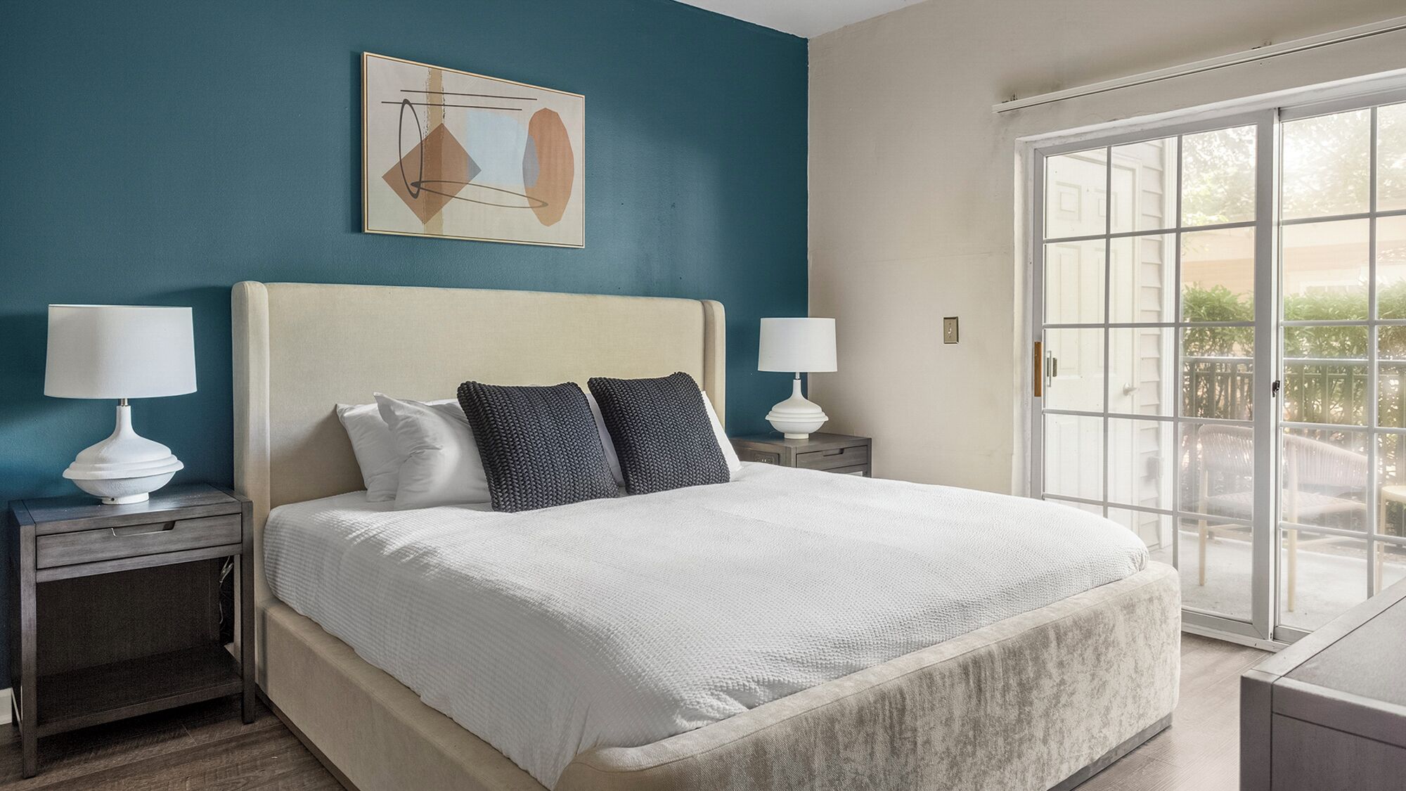 Une chambre avec un grand lit, deux tables de chevet avec des lampes, un tableau au mur et une grande fenêtre donnant sur l’extérieur.