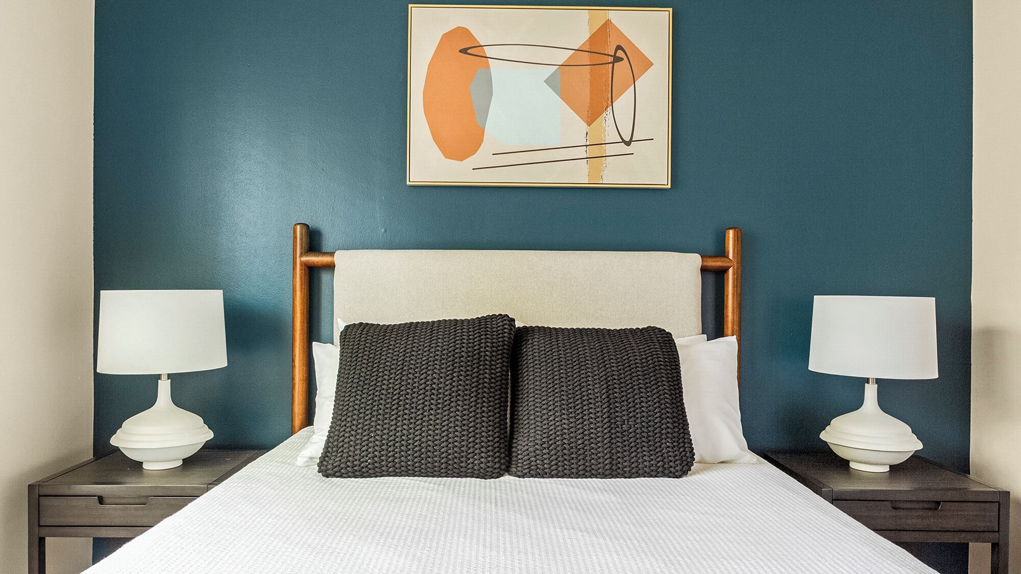 A neatly made bed with a white comforter and two pillows, flanked by two bedside tables with white lamps and a white bowl.