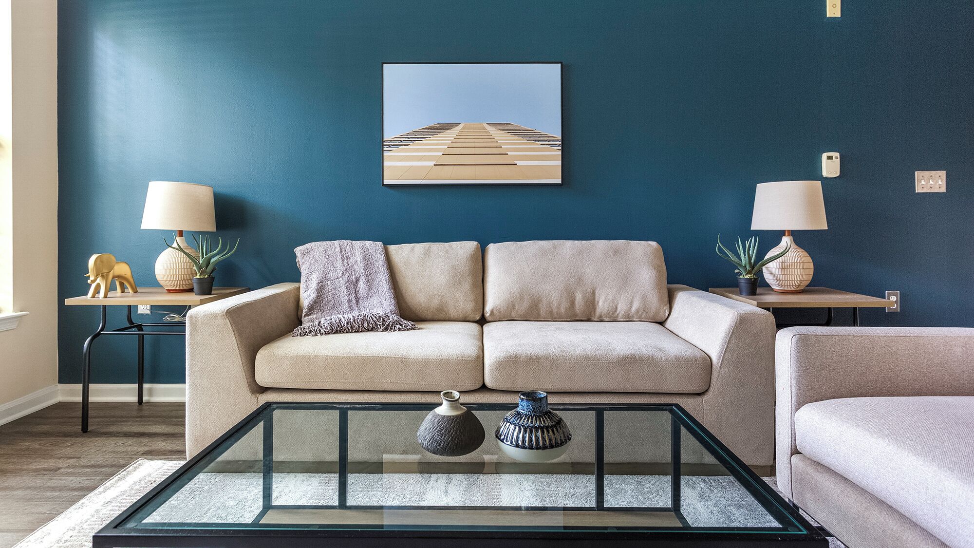 Ein Wohnzimmer mit einem beigefarbenen Sofa, einem Glastisch, zwei Beistelltischen mit Lampen und einem gerahmten Bild an der Wand.
