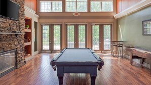 Un vaste espace intérieur comprenant une table de billard, une cheminée en pierre et de grandes fenêtres laissant entrer la lumière naturelle.