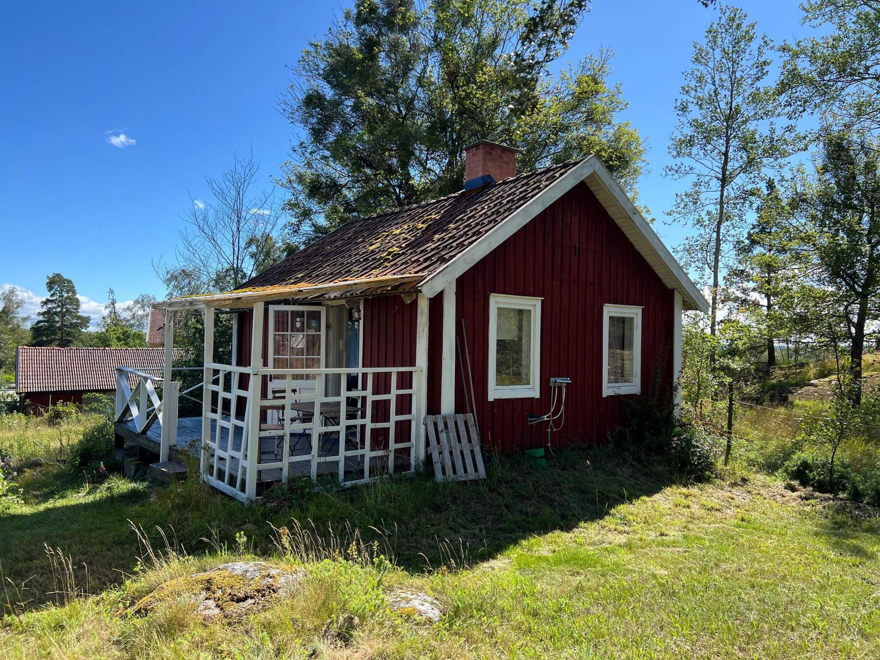 A red wooden cabin with a porch, surrounded by trees and grass.