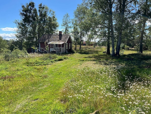 Une maison en bois rouge, dotée d’un porche, entourée d’arbres et d’un champ verdoyant.