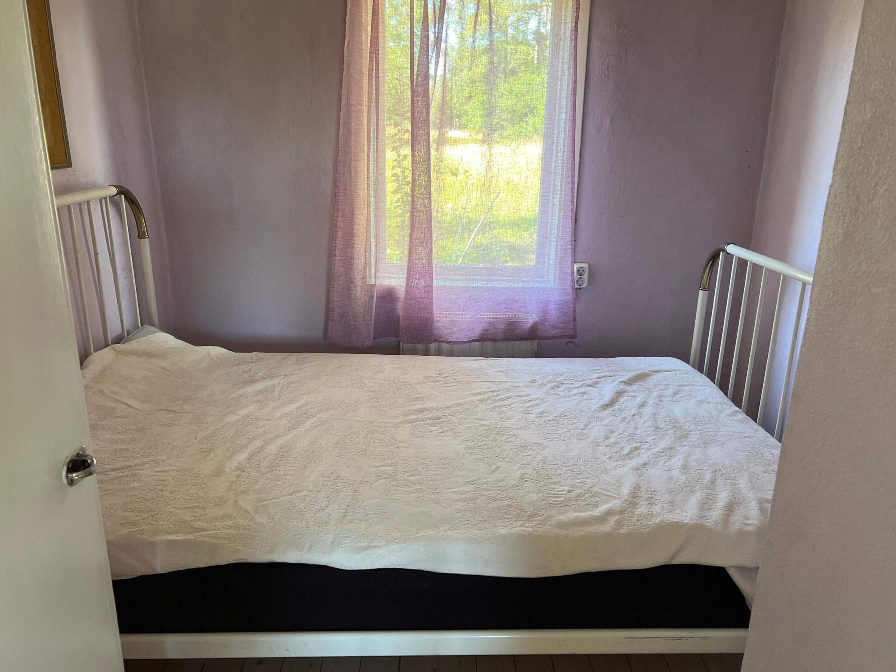 A small, single-bed room with a white mattress, a window with sheer curtains, and a white radiator.