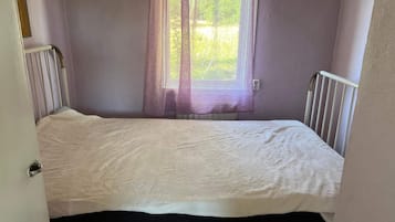 A small, single-bed room with a white mattress, a window with sheer curtains, and a white radiator.