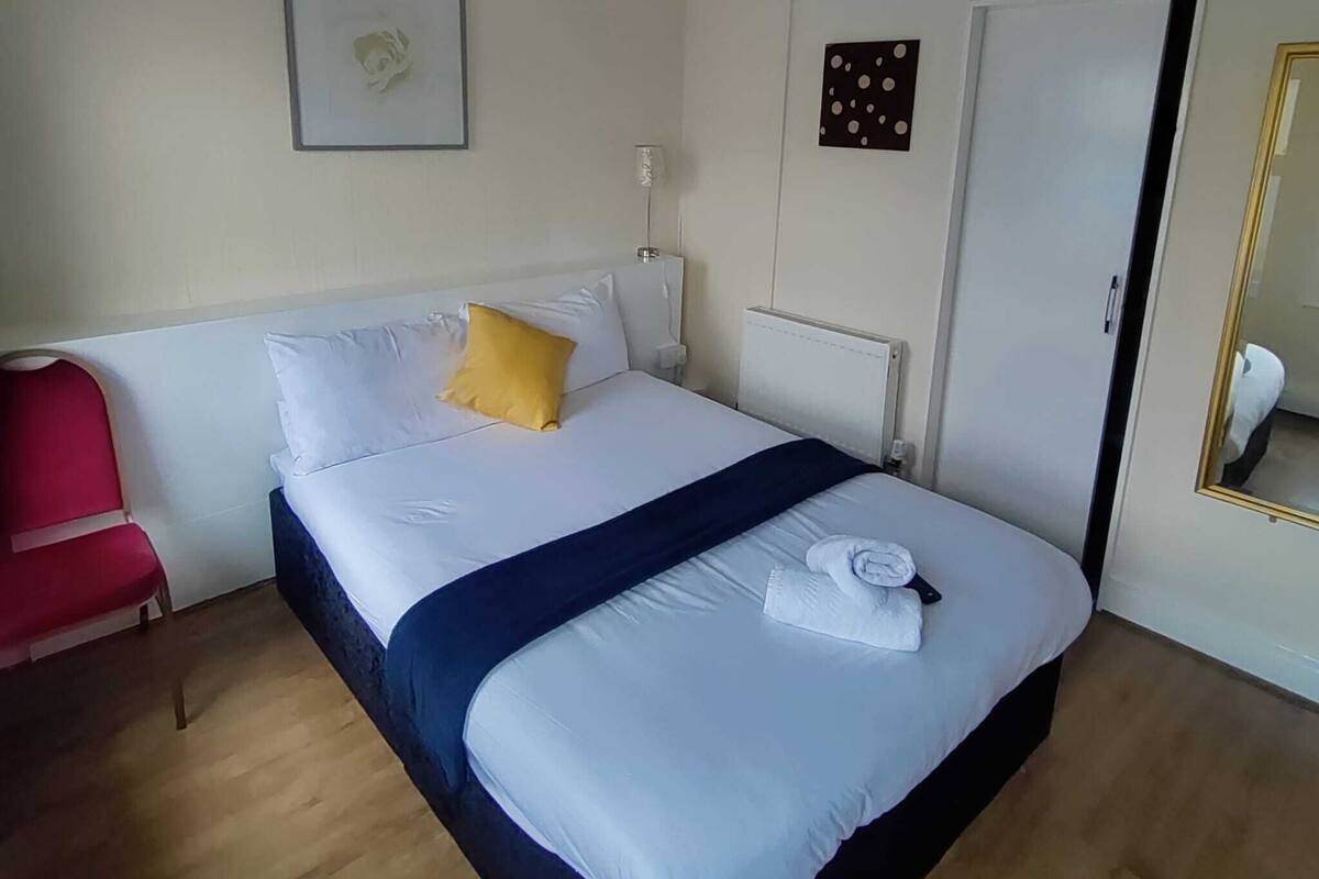 Une chambre d’hôtel avec un lit, une chaise rouge, un tableau encadré au mur et une porte avec un miroir.