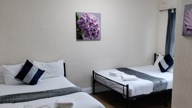 Une chambre d’hôtel avec deux lits, du linge de lit blanc, une couvre-lit grise à motifs et une seule œuvre d’art représentant des fleurs violettes accrochée au mur.