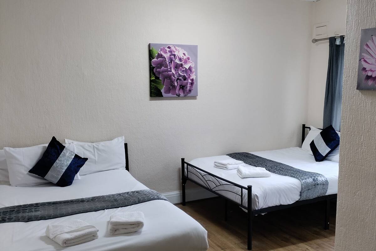 Une chambre d’hôtel avec deux lits, du linge de lit blanc, une couvre-lit grise à motifs et une seule œuvre d’art représentant des fleurs violettes accrochée au mur.