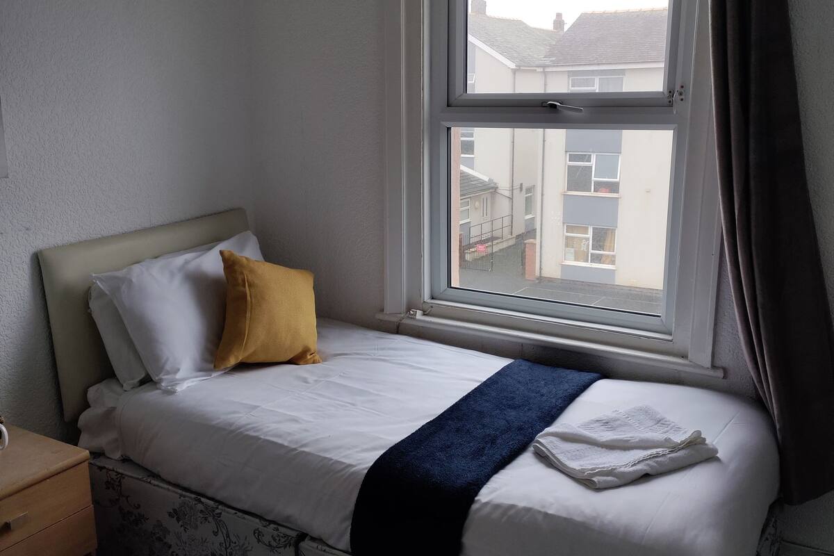 Un lit simple avec une tête de lit, un oreiller jaune, un oreiller blanc, une couverture bleue et une serviette blanche. Il y a une fenêtre avec des rideaux, une table de chevet en bois avec une lampe et un couvre-matelas à motifs floraux.