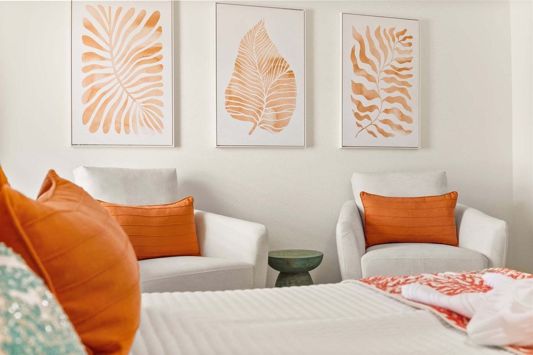 A room with two white armchairs, orange cushions, a green side table, and a bed with a white and red patterned blanket.