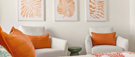 A room with two white armchairs, orange cushions, a green side table, and a bed with a white and red patterned blanket.