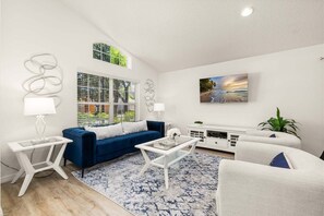 A modern living room with a blue sofa, white coffee table, and a large window with blinds.