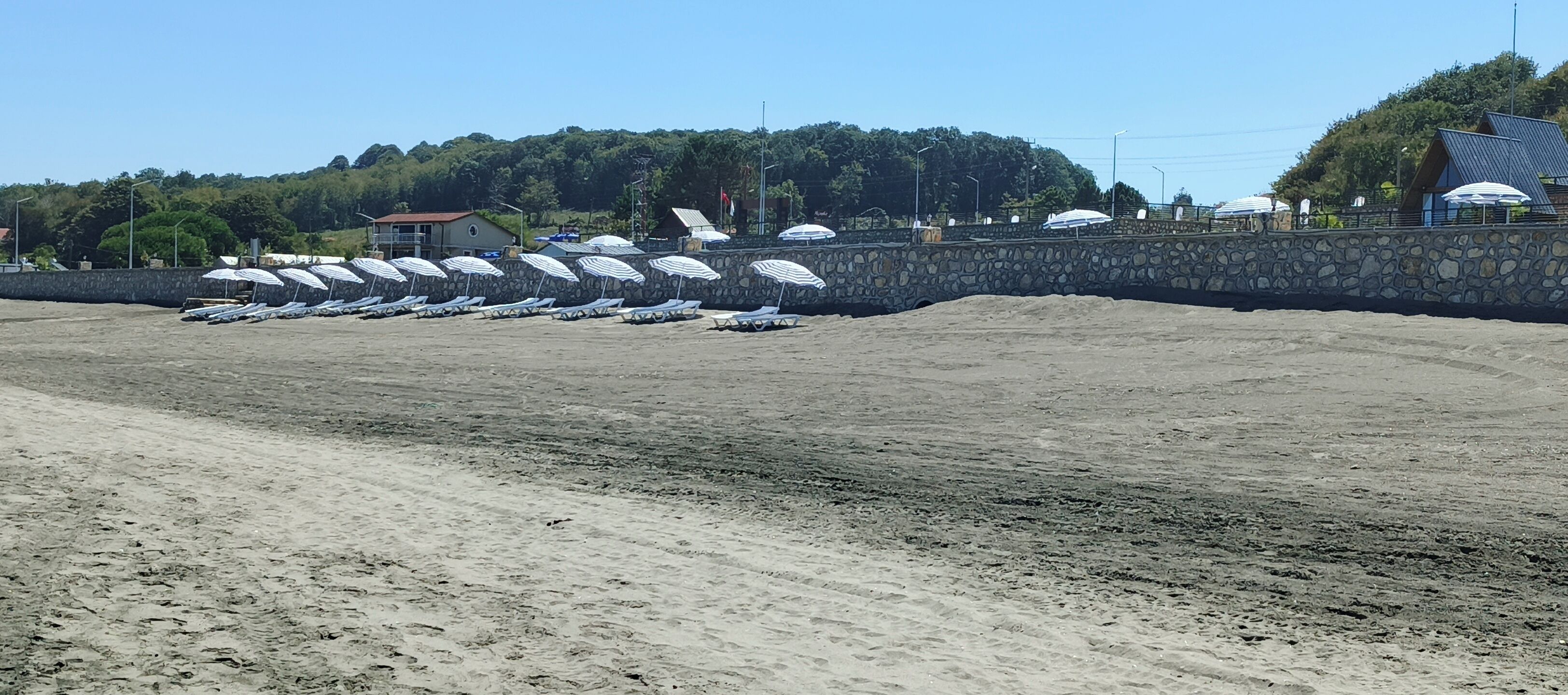 Une plage de sable avec des rangées de chaises longues et de parasols, un mur de pierre et des bâtiments en arrière-plan.