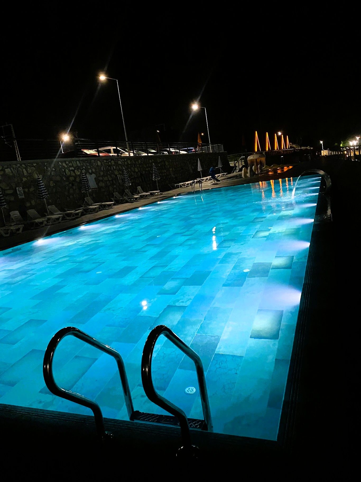 A swimming pool with clear blue water, surrounded by a stone wall and equipped with lounge chairs and umbrellas.
