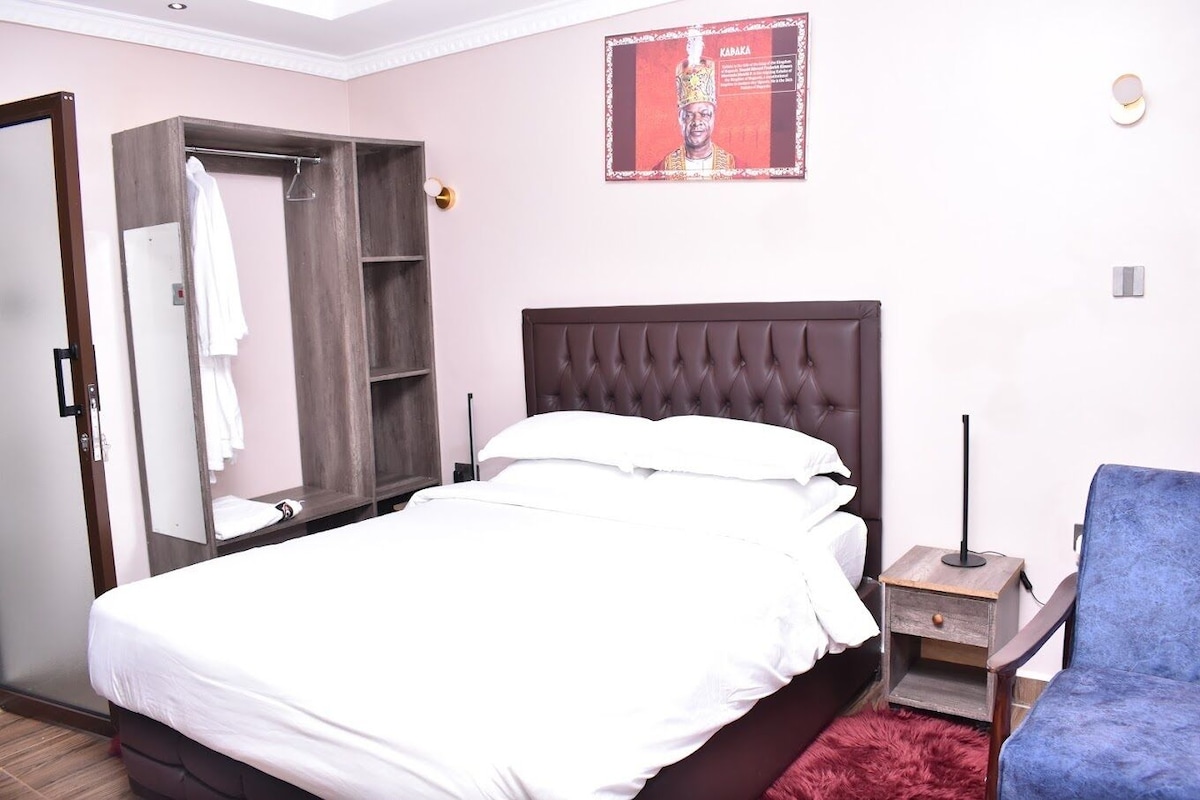 A neatly made bed with white linens, a wooden wardrobe, a blue chair, and a small wooden nightstand with a lamp.