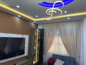 A modern living room with a flat-screen TV, a blue sofa, and a decorative ceiling light fixture. - Luxury One bedroom Apartment R+1 (Maritime Region)