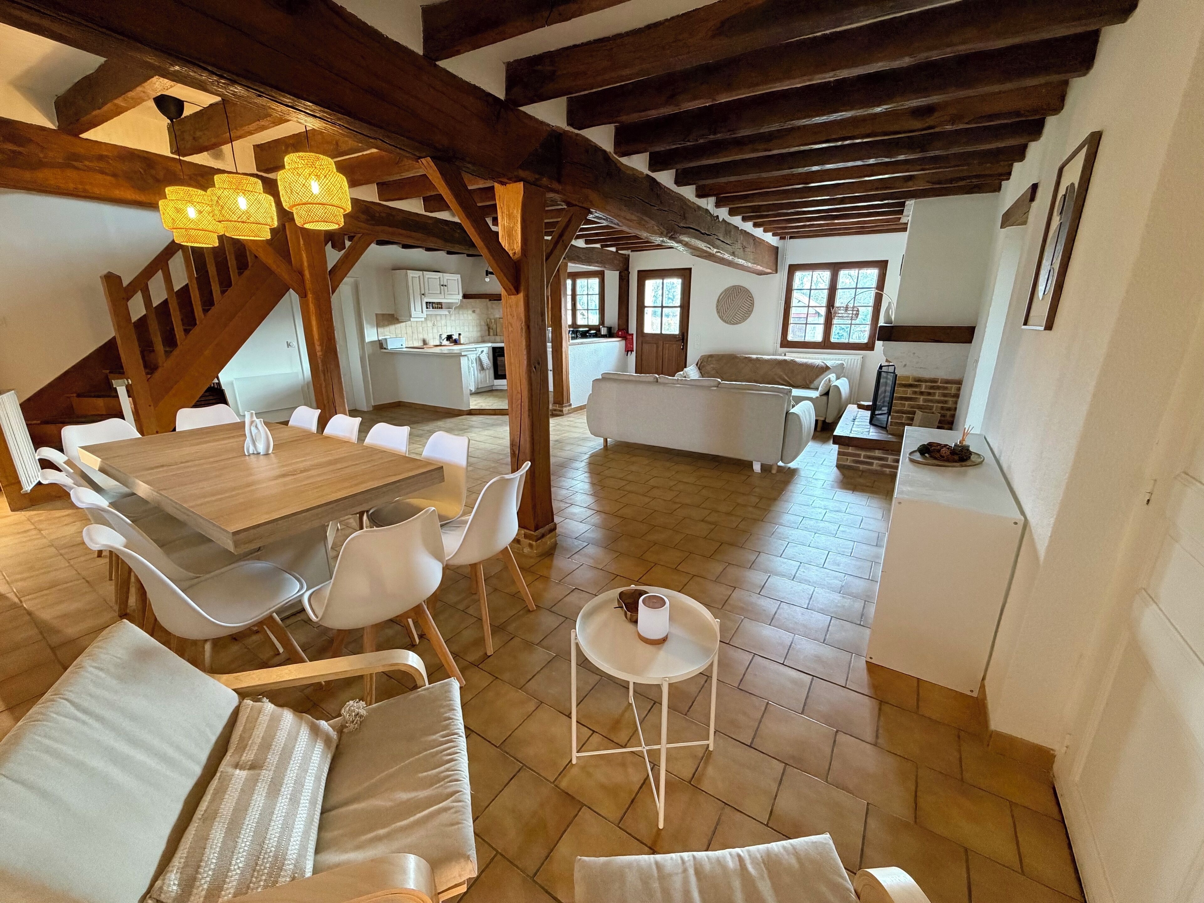 A spacious dining area with a wooden table and white chairs, a cozy seating area with a sofa, and a kitchen in the background.