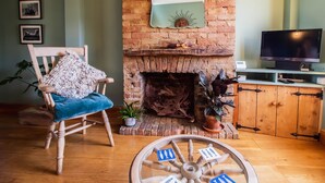A cozy living room with a brick fireplace, a wooden chair with a blue cushion, a television on a wooden cabinet, and a decorative wheel on the floor. - Sun House Apartment Seaford (Seaford)
