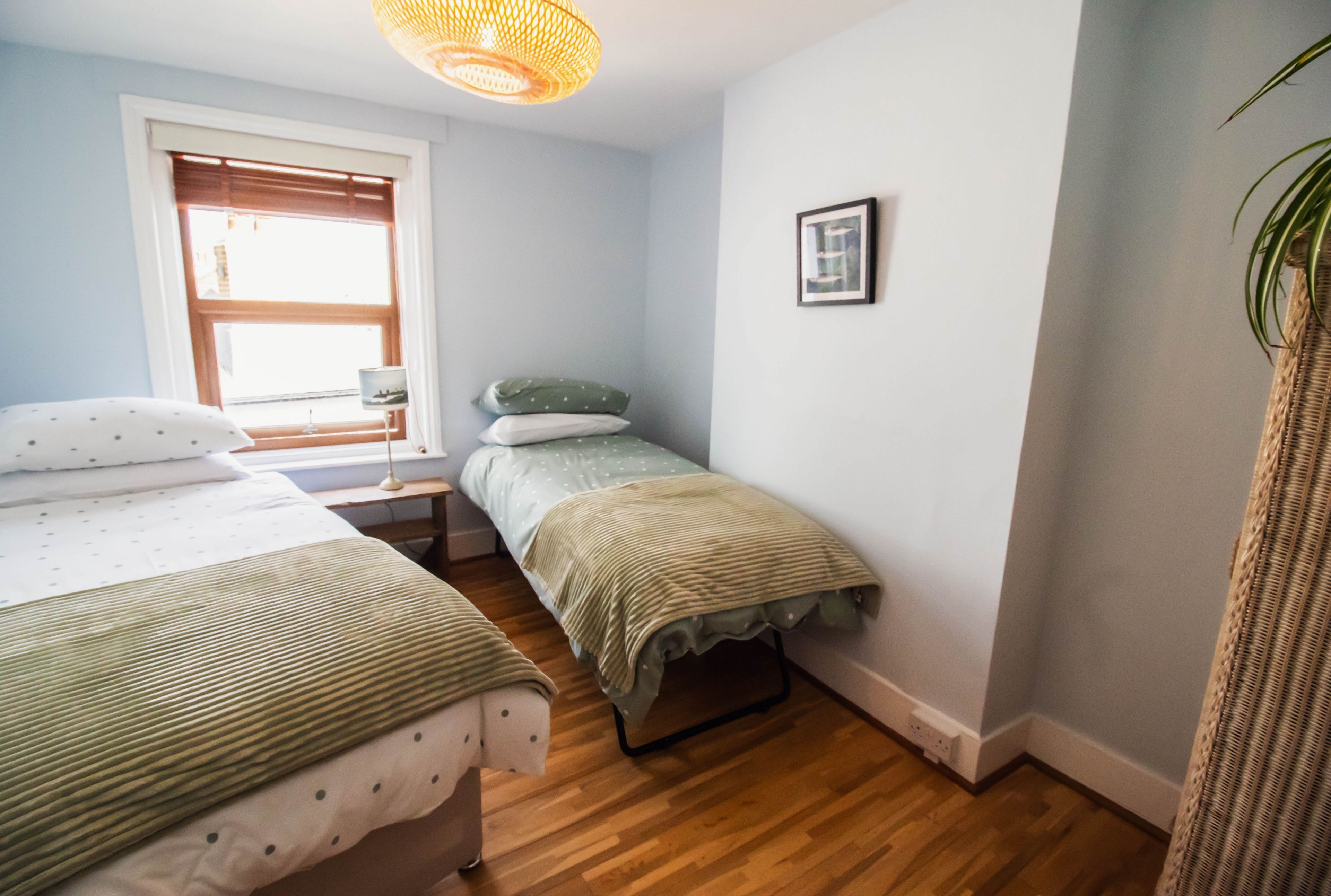 A bedroom with two beds, a window with blinds, a bedside table with a lamp, and a framed picture on the wall.