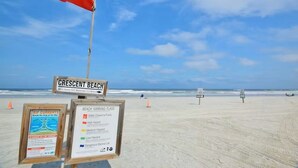 A beach with a red flag indicating a rip current warning, a sign for Crescent Beach, and beach warning flags detailing water conditions. - Cottage & Bungalow | Coastal 2-Unit Retreat (St. Augustine)