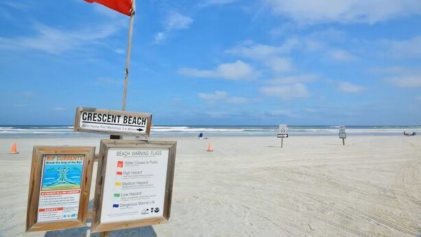 A beach with a red flag indicating a rip current warning, a sign for Crescent Beach, and beach warning flags detailing water conditions.