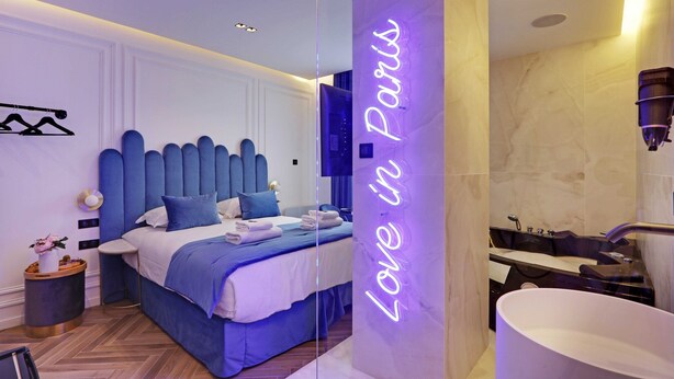 A modern bedroom with a large bed, a bedside table with a vase, and a neon sign reading "Love in Paris".