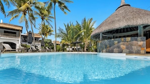 A swimming pool with lounge chairs and umbrellas, surrounded by palm trees and a thatched-roof structure.