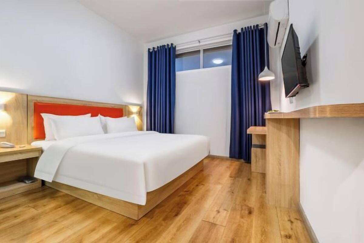 Ein Hotelzimmer mit einem Bett, Holzboden, einem an der Wand montierten Fernseher und einem Nachttisch mit Lampe.