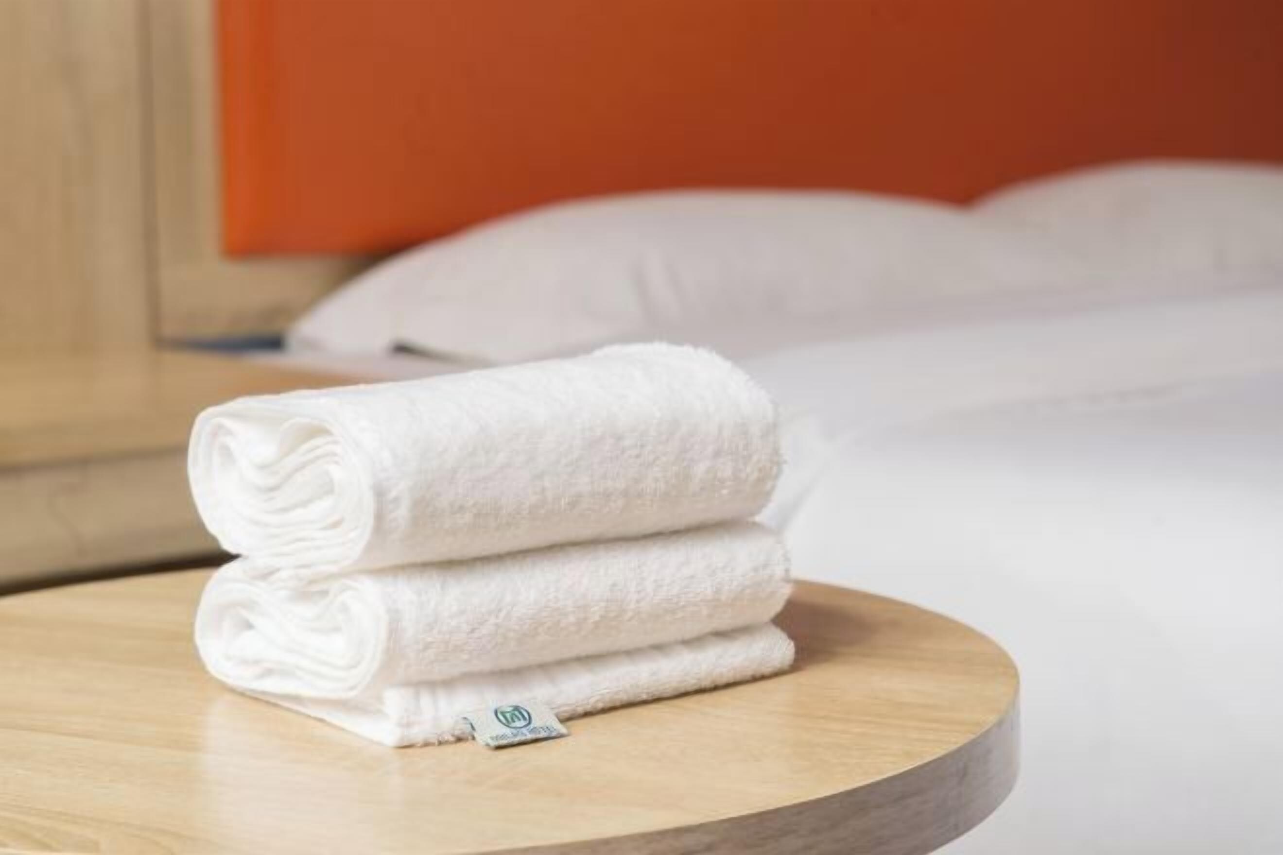 A stack of white towels on a wooden table in a room with a bed and an orange wall in the background.