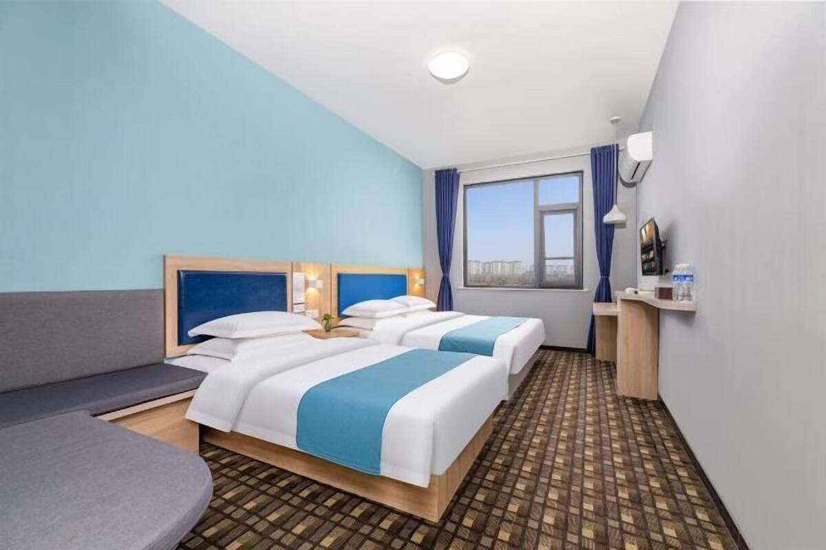 Ein Hotelzimmer mit zwei Betten, einem Schlafsofa, einem Fenster mit Blick auf die Stadt und einem kleinen Schreibtisch mit Lampe.