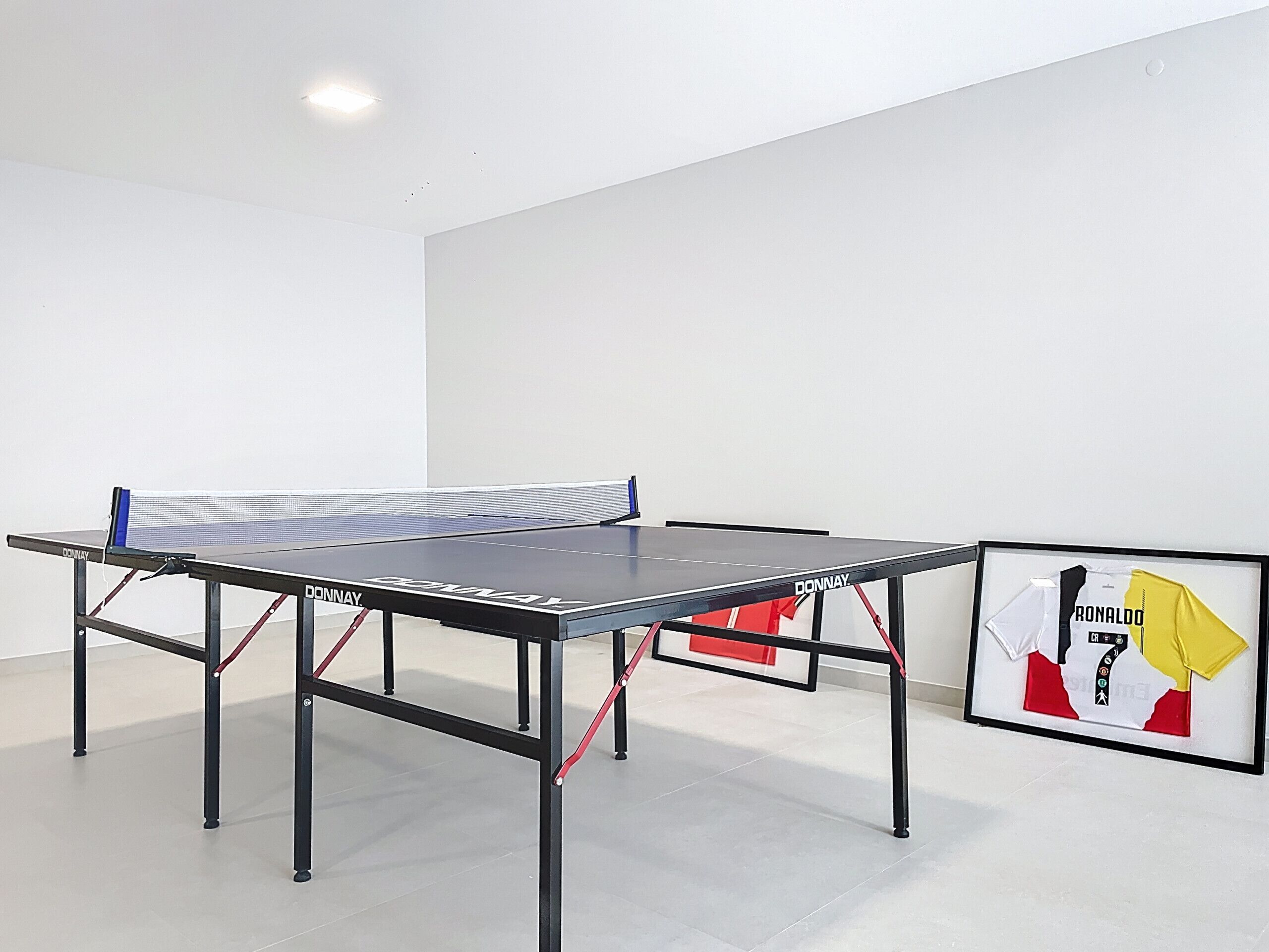Una mesa de ping pong con red, con la marca 'DONNAY', en una habitación con paredes blancas y un cuadro con una camiseta deportiva.