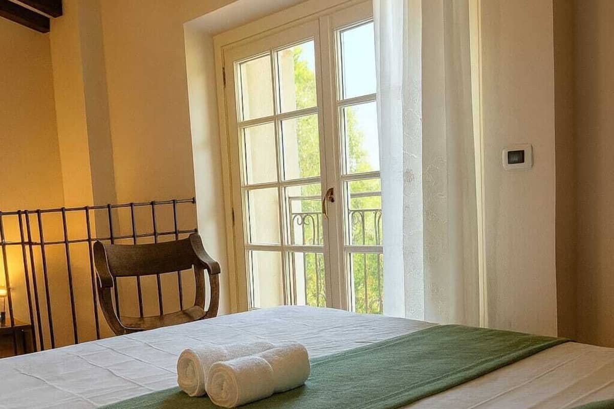 A hotel room with a bed, a window with curtains, a chair, and a view of greenery outside.