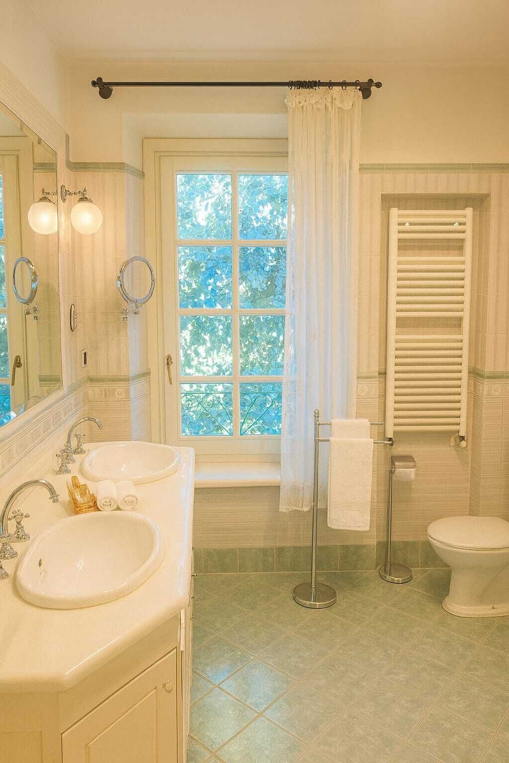 Un baño con dos lavamanos, inodoro, ducha con puerta de cristal y ventana con vistas a árboles.