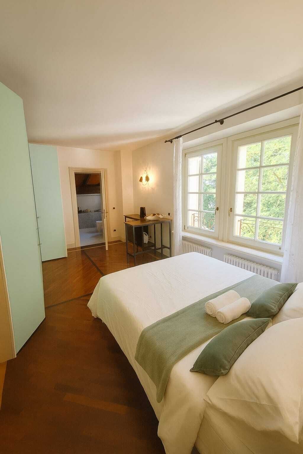 A bedroom with a large bed, a desk, and a window with a view of greenery.